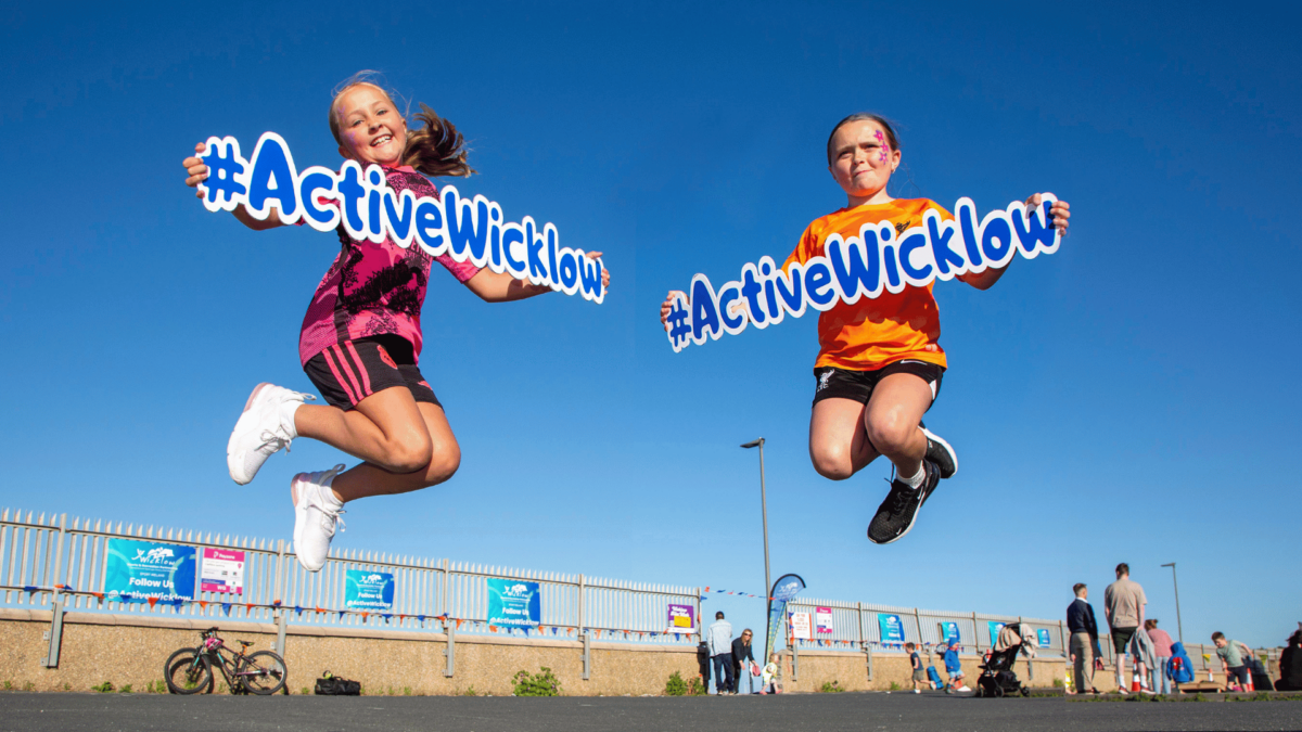 Two young girls jumping in the air, smiling and holding #ActiveWicklow signs against a clear blue sky at an outdoor event, with people, bicycles, and event posters visible in the background.
