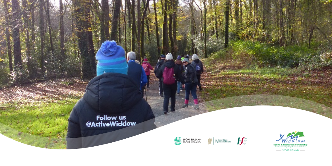 Wide banner image of a group walking away from the camera down a gravel path surrounded by autumn trees. In the foreground, a person wears a blue beanie and a coat with the text "Follow us @ActiveWicklow". The bottom of the image is overlaid with a white wavy graphic showcasing partner logos from left to right: Spórt Éireann / Sport Ireland, An Roinn Sláinte / Department of Health, HSE, and Wicklow Sports & Recreation Partnership.
